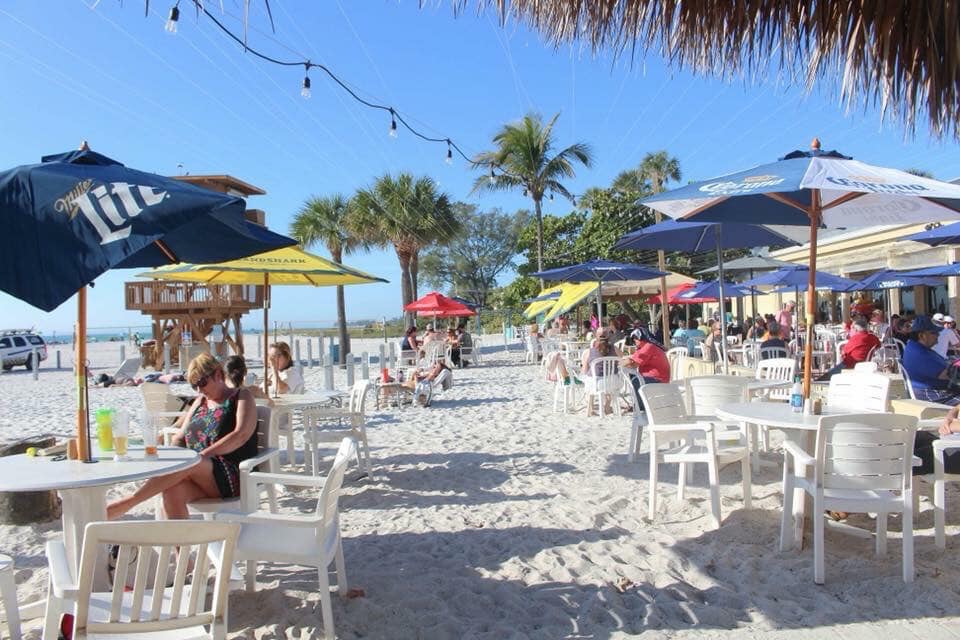 Rows of beach umbrellas and tables in the sand with palm trees and the Gulf behind