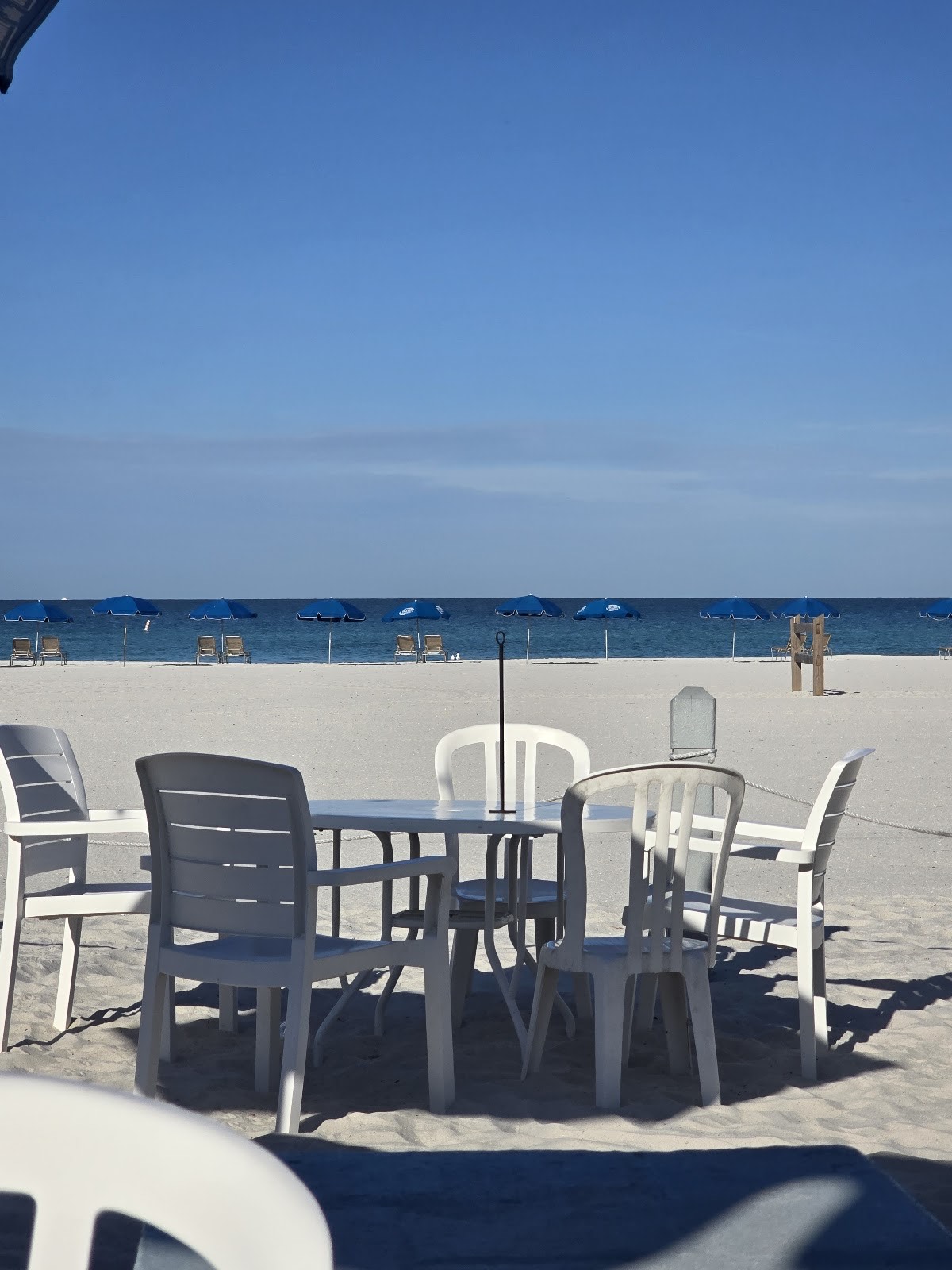 Beach table and chairs in the white sand looking out to blue Gulf umbrellas and water