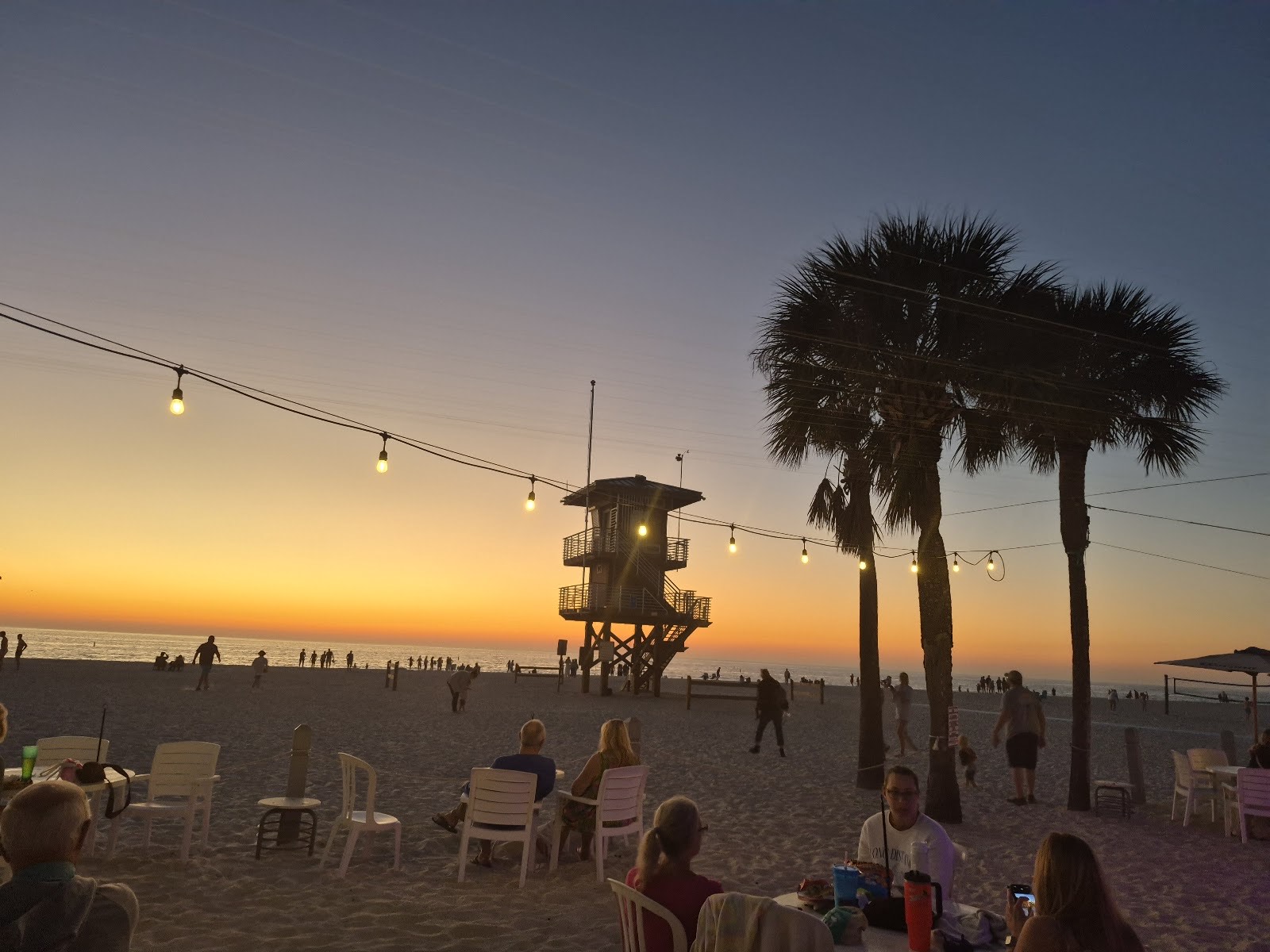 Sunset over Manatee Beach with the lifeguard tower, palms and string lights above guests